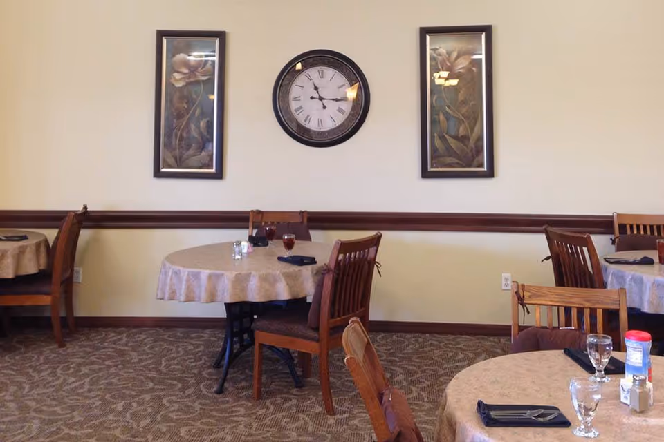 Interior view of a senior living community dining room with round tables covered in beige tablecloths, wooden chairs, a wall clock showing 3:45, and two framed floral paintings on the wall.