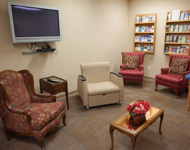 Small sitting room with upholstered chairs, a wall-mounted TV, brochure racks, and a coffee table with flowers.