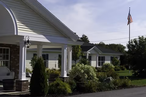 Covered entrance and single-story residential buildings surrounded by landscaped shrubs and an American flag.