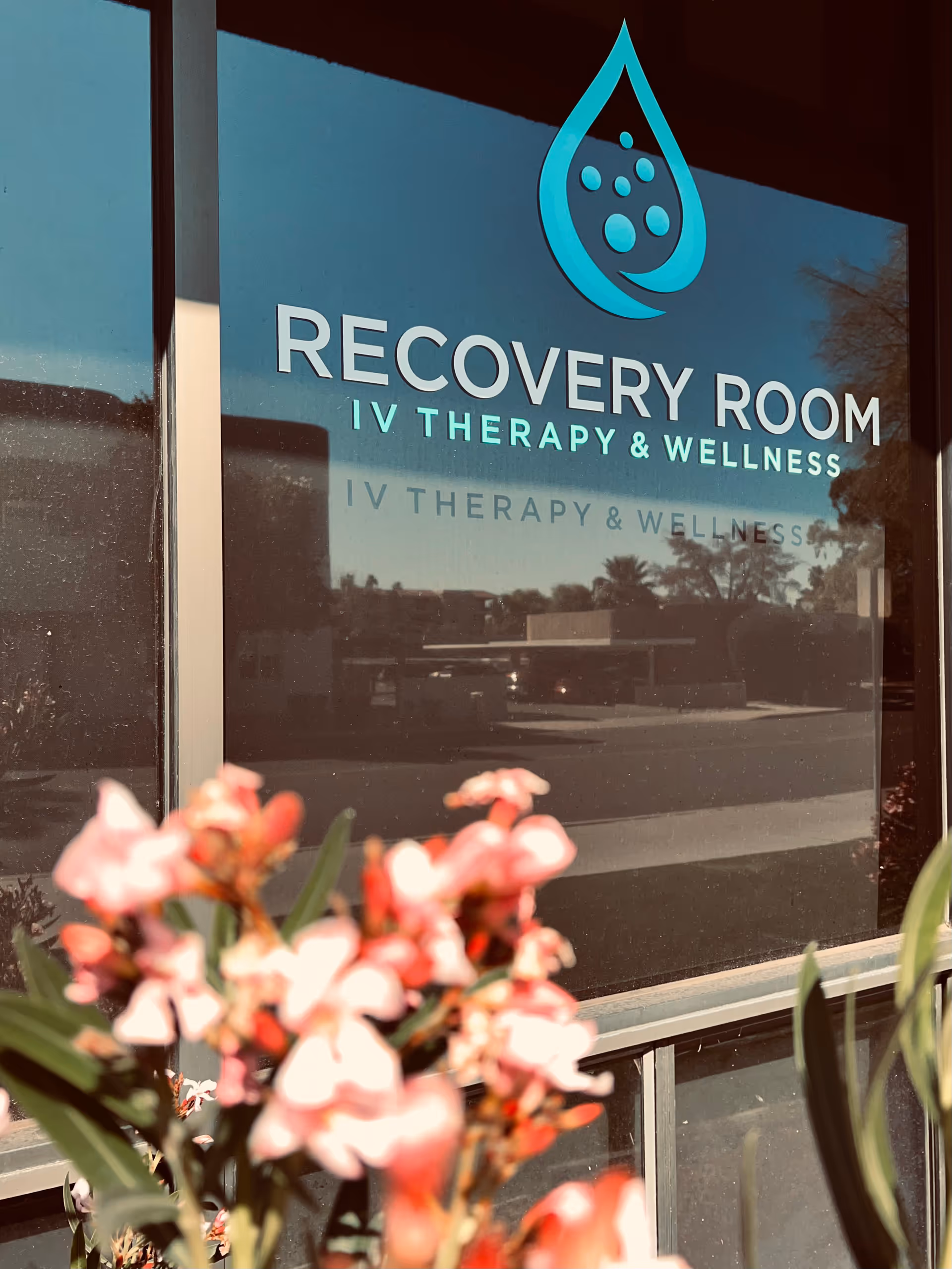 Glass storefront sign reads Recovery Room IV Therapy and Wellness with pink flowers in foreground