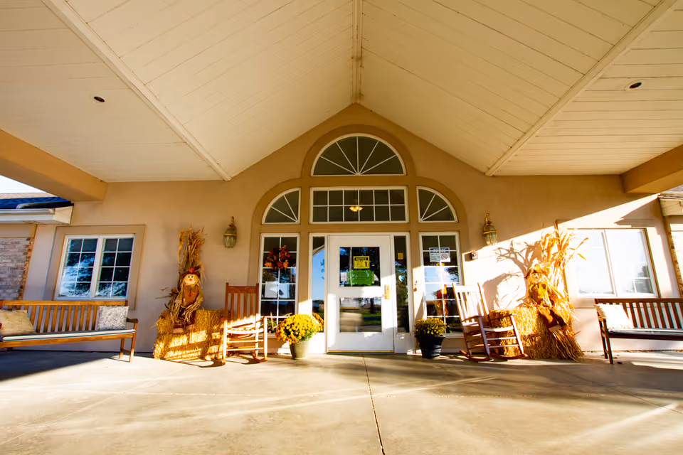 Entrance of a building with a covered porch featuring two wooden benches and two rocking chairs. There are autumn-themed decorations including scarecrows made of straw and potted yellow flowers near the glass double doors with arched windows above.