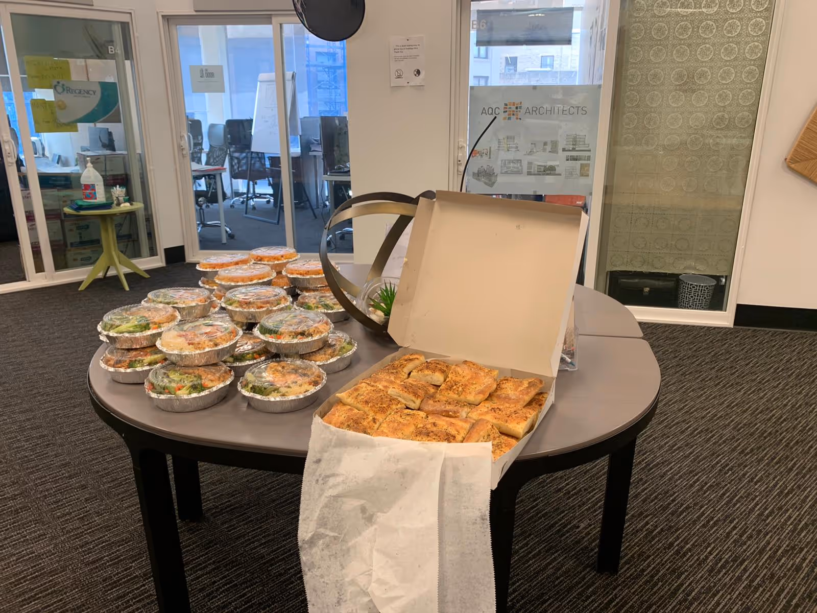 Table with multiple aluminum trays of food and pastries indoors, in a conference area.