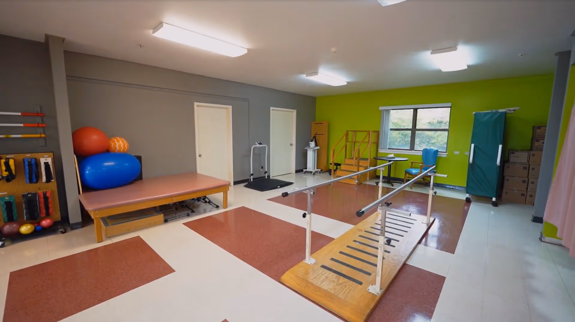 Physical therapy room with parallel walking bars, a treatment table, exercise balls, and other rehab equipment.