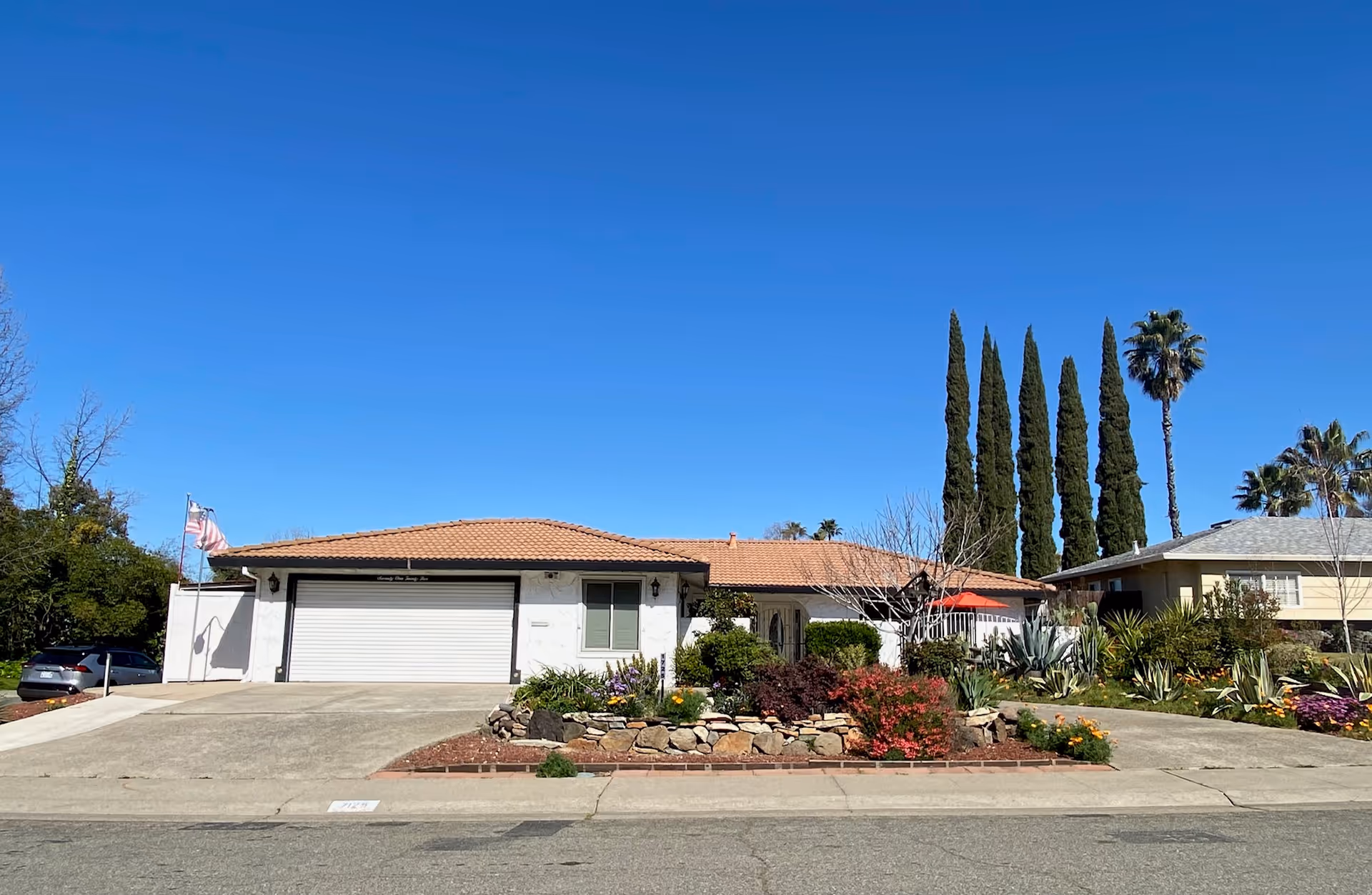 Single-story stucco house with a tiled roof, two-car garage and landscaped front yard with tall cypress trees under a clear blue sky.
