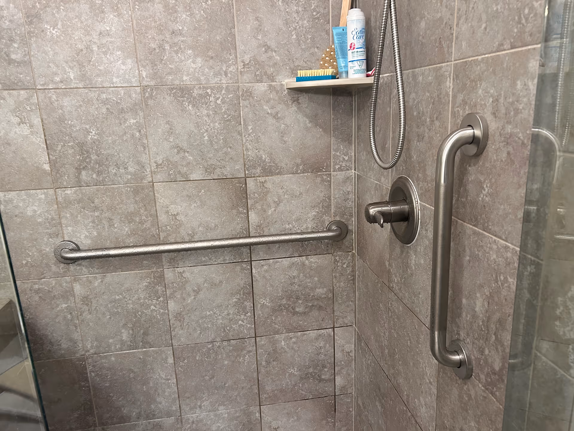 Grey tiled walk-in shower with metal grab bars and toiletries on a shelf.