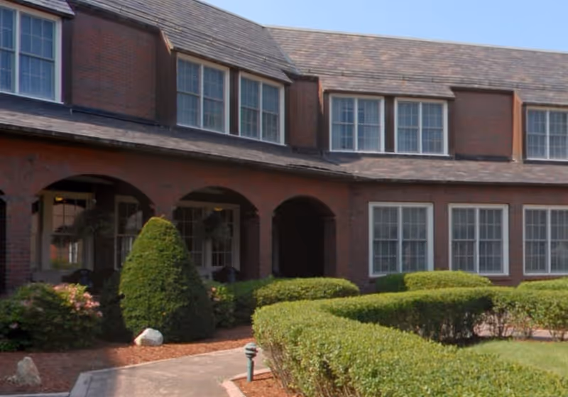 Exterior view of a brick building with multiple windows and a shingled roof. The building features arched entryways and is surrounded by neatly trimmed bushes and landscaped greenery under a clear blue sky.