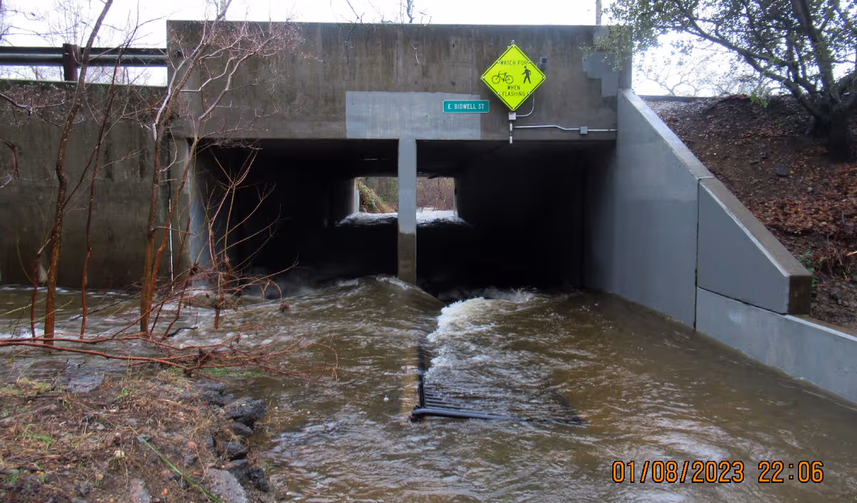 Flooded concrete underpass with rushing water, railing, and stairs beside a flood control channel.