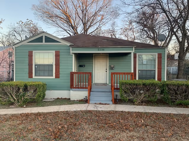 Small green single-story house with a front porch, steps, red shutters, and trimmed bushes.