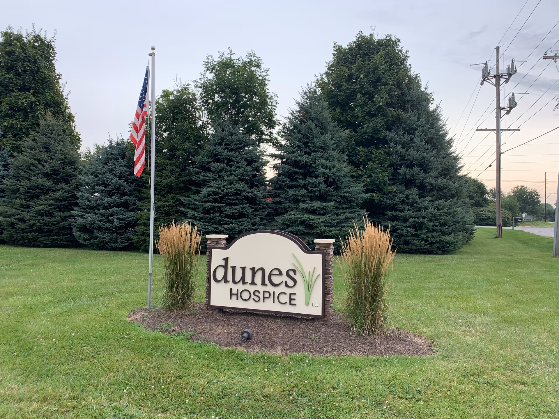 Dunes Hospice sign on a grassy lawn with tall evergreens and an American flag nearby.