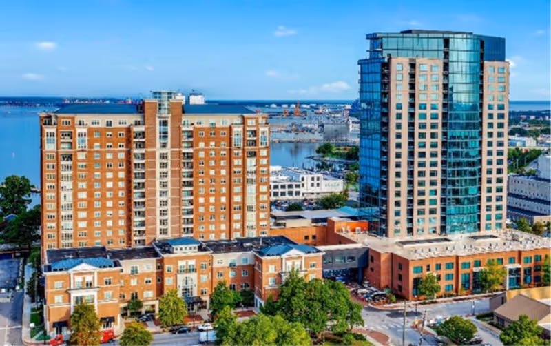 Aerial view of Harbor’s Edge facility showing multiple connected buildings including a tall glass tower and several brick buildings, with a waterfront and harbor in the background under a clear blue sky.