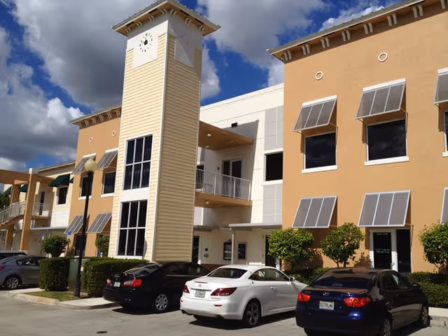 Exterior view of a tan multi-story senior living building with a clock tower and parked cars.