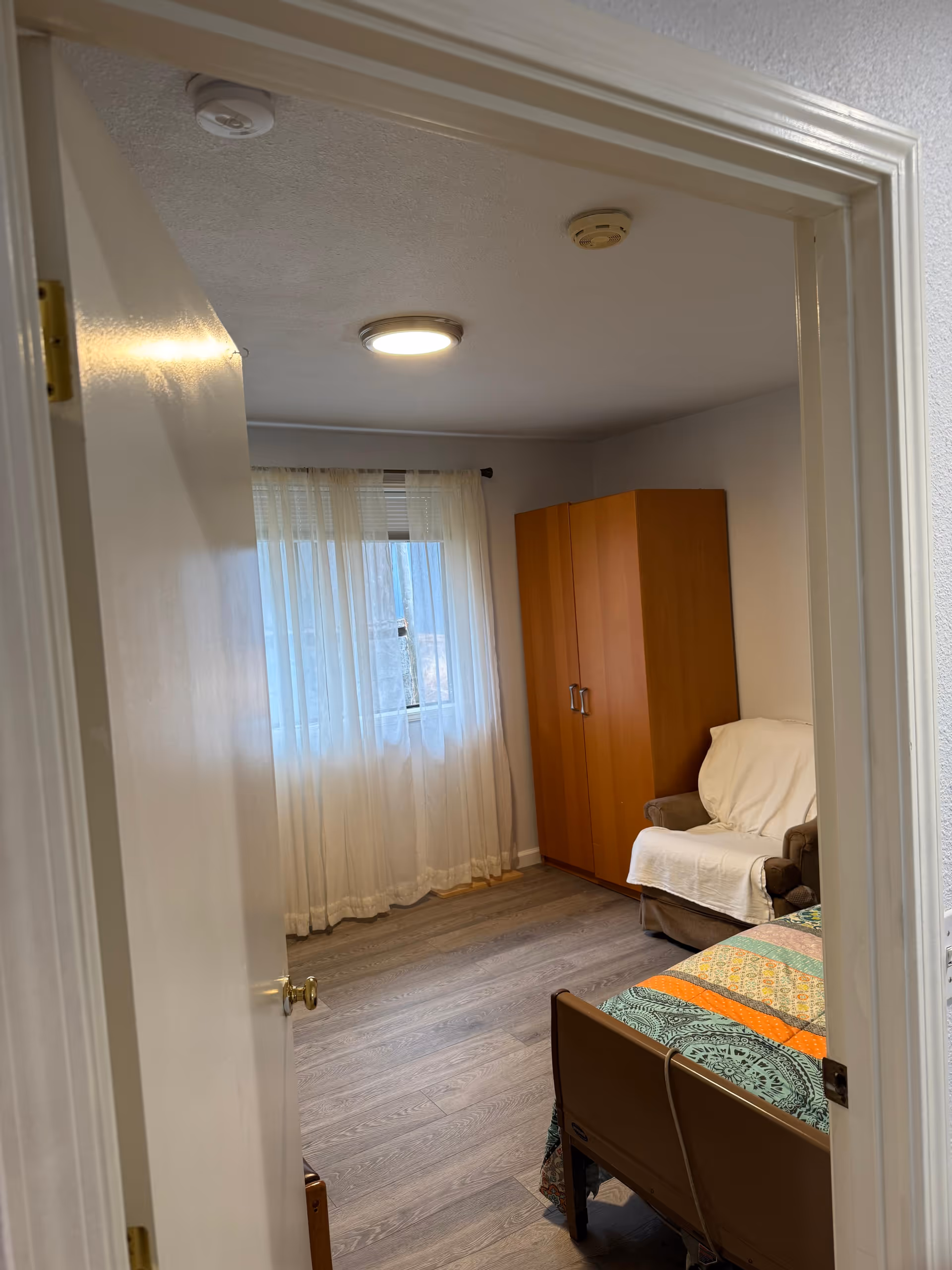 Small bedroom in a care facility with a bed, wardrobe, chair, and sheer curtains.