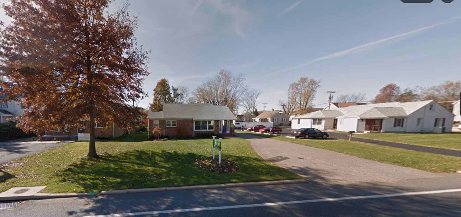 A suburban street with a single-story brick building center, green lawn, and parked cars.