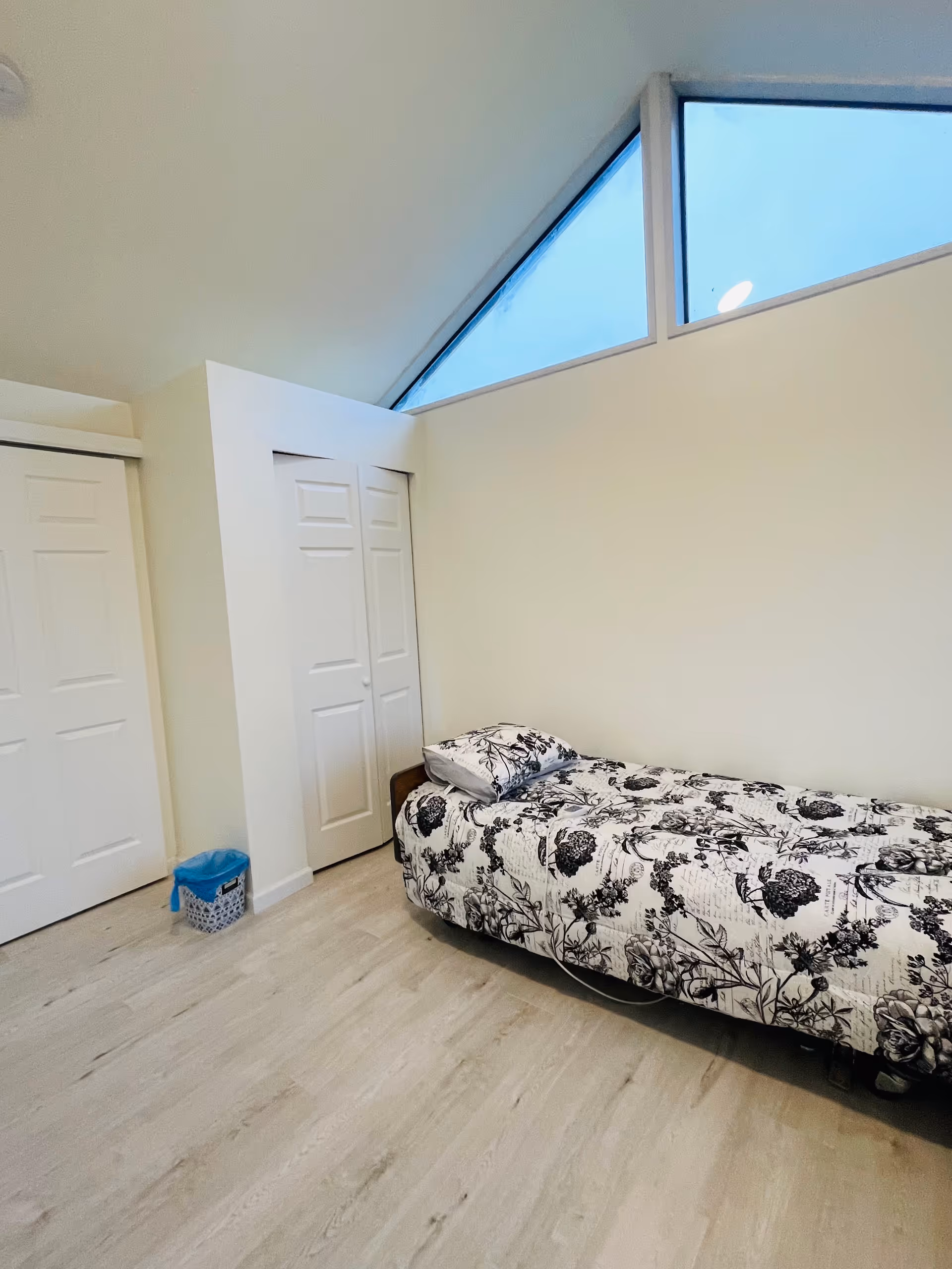 A minimal bedroom with a single floral bed, closets, and a high triangular window.