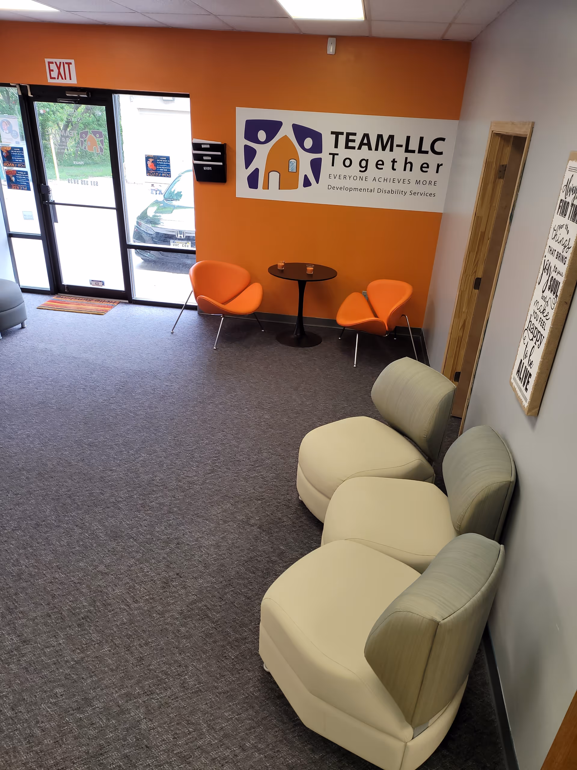 Orange-walled lobby with orange chairs, cream seating, round table, and TEAM-LLC sign near exit.