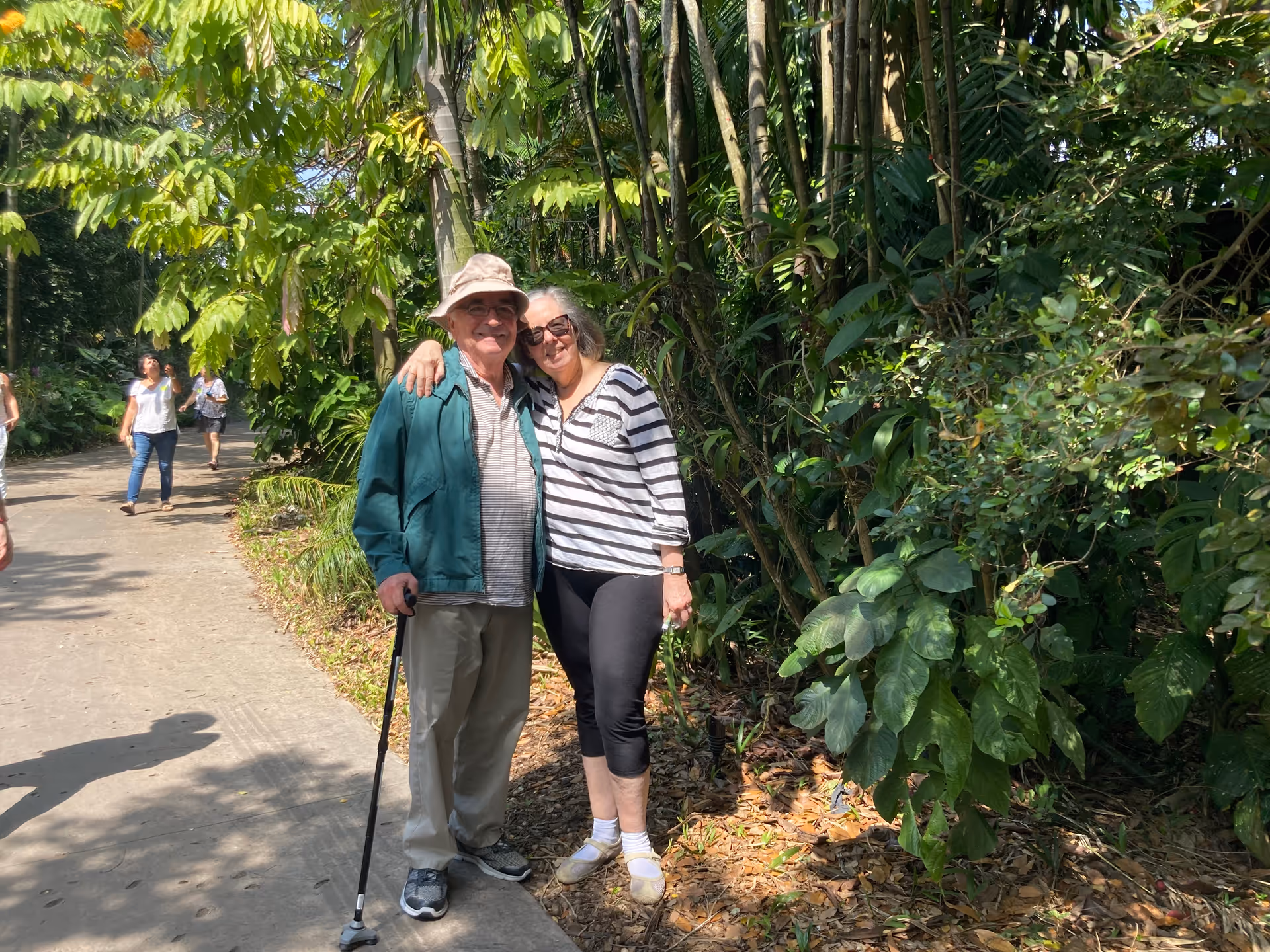 An elderly man and woman standing on a paved path surrounded by lush green foliage. The man is wearing a hat, glasses, a green jacket, and holding a cane, while the woman is wearing sunglasses, a striped shirt, and black pants. They are smiling and posing closely together with the woman's arm around the man's shoulder. Other people are walking in the background along the path.
