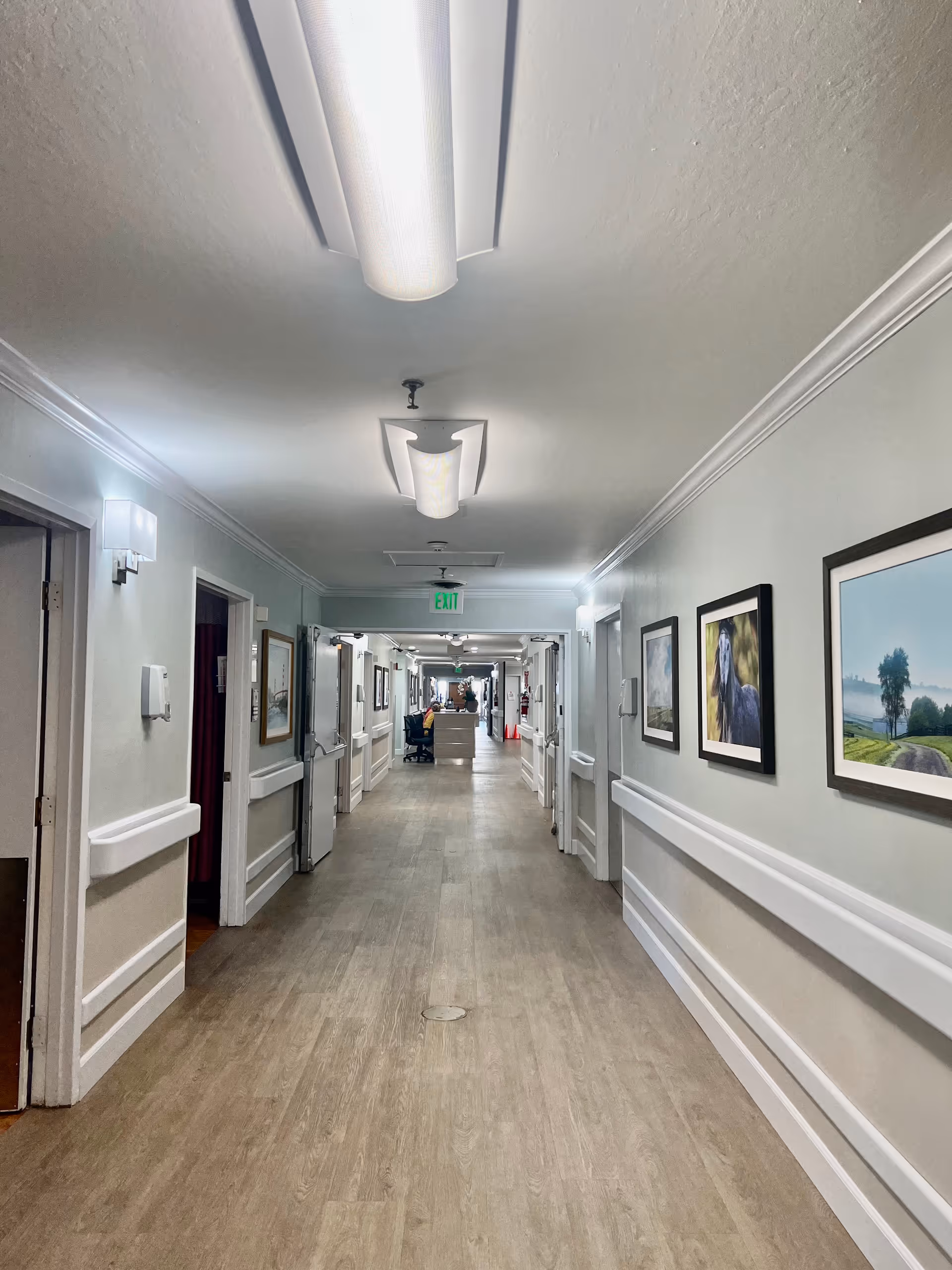 Long hallway inside a senior living facility with framed pictures and ceiling lights.