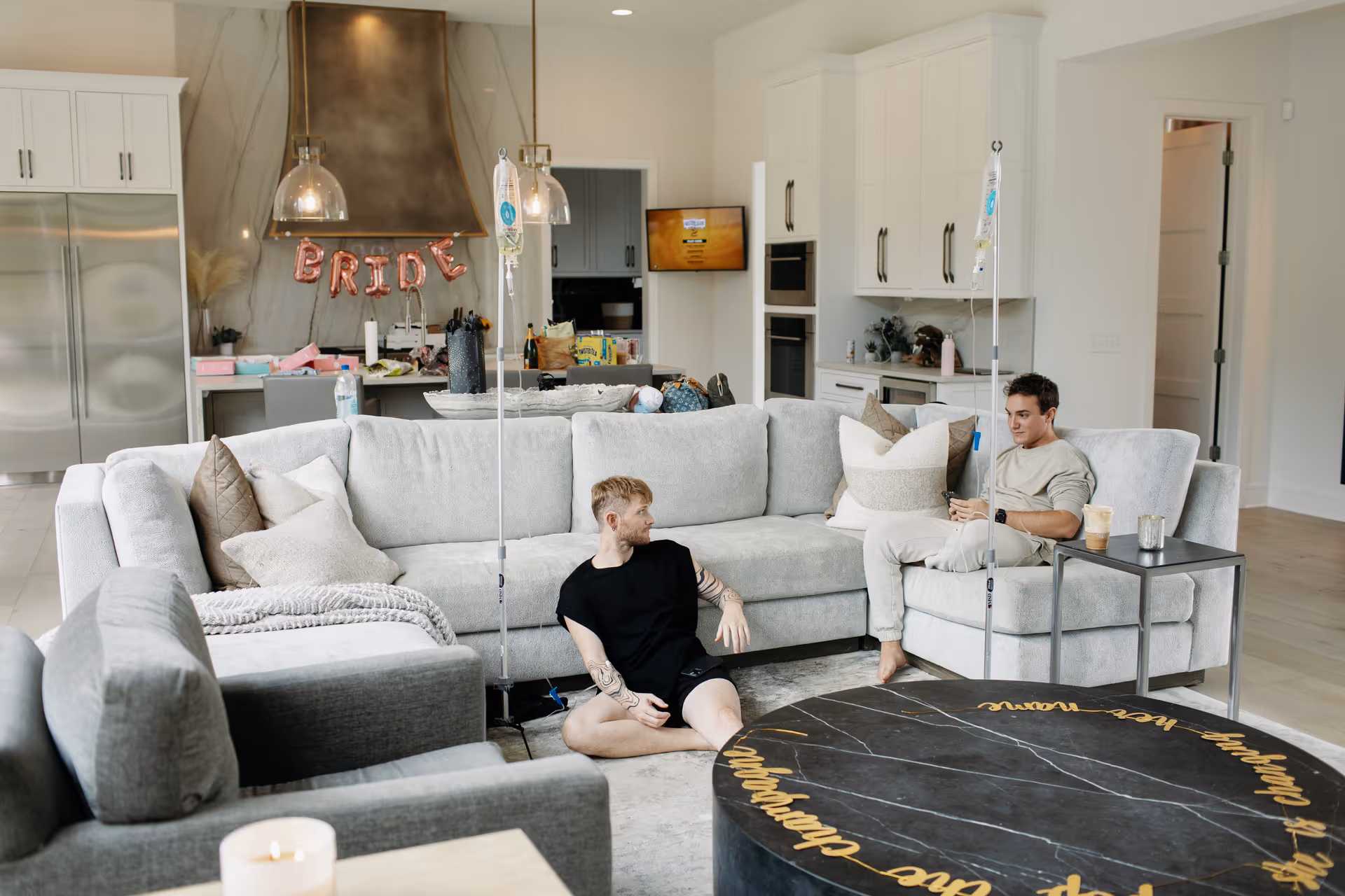 Two men sit on a gray sectional in an open concept living room with a kitchen.