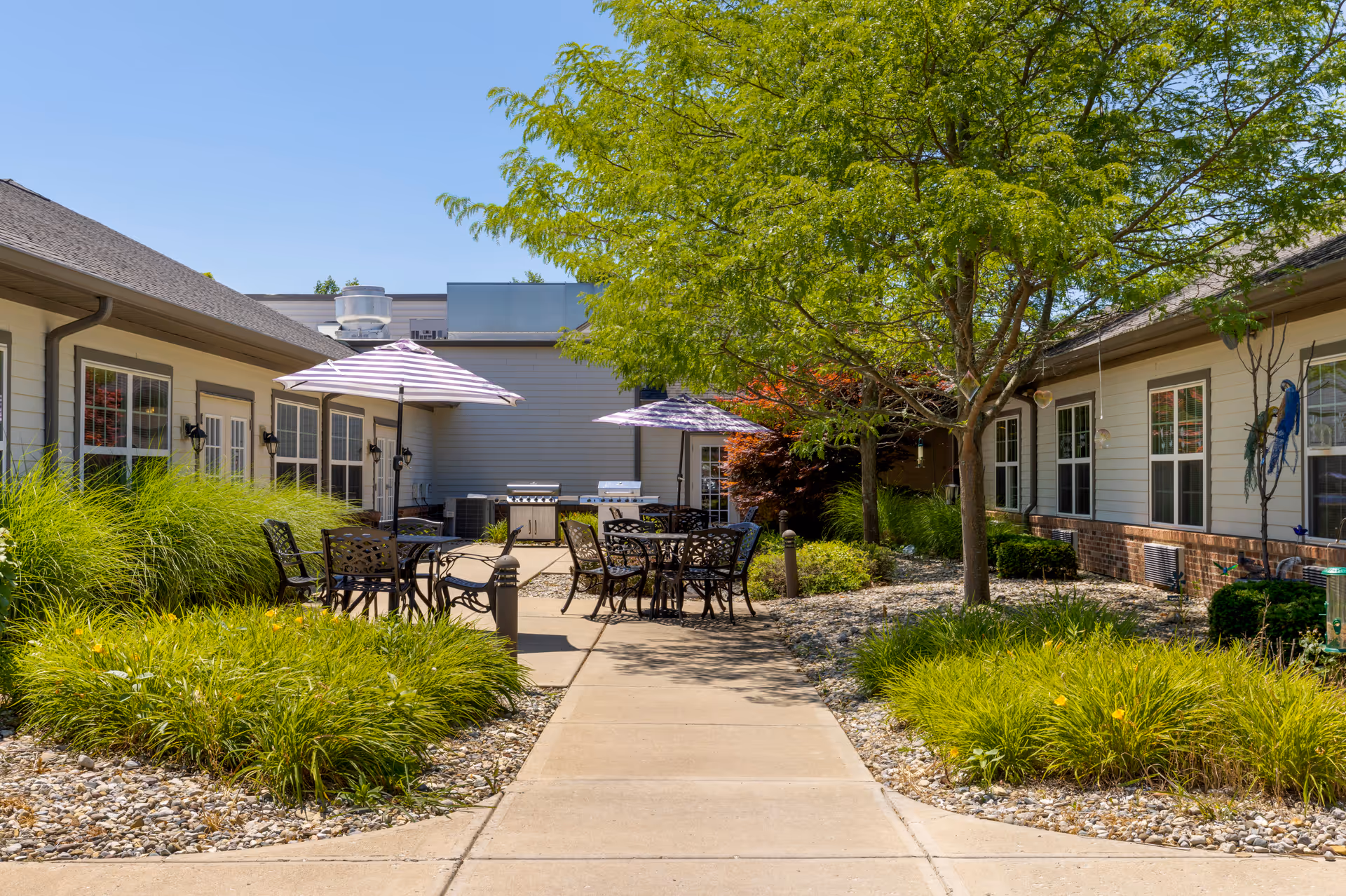 Sunlit courtyard with patio tables, umbrellas, chairs, trees and landscaped paths between single-story building wings.