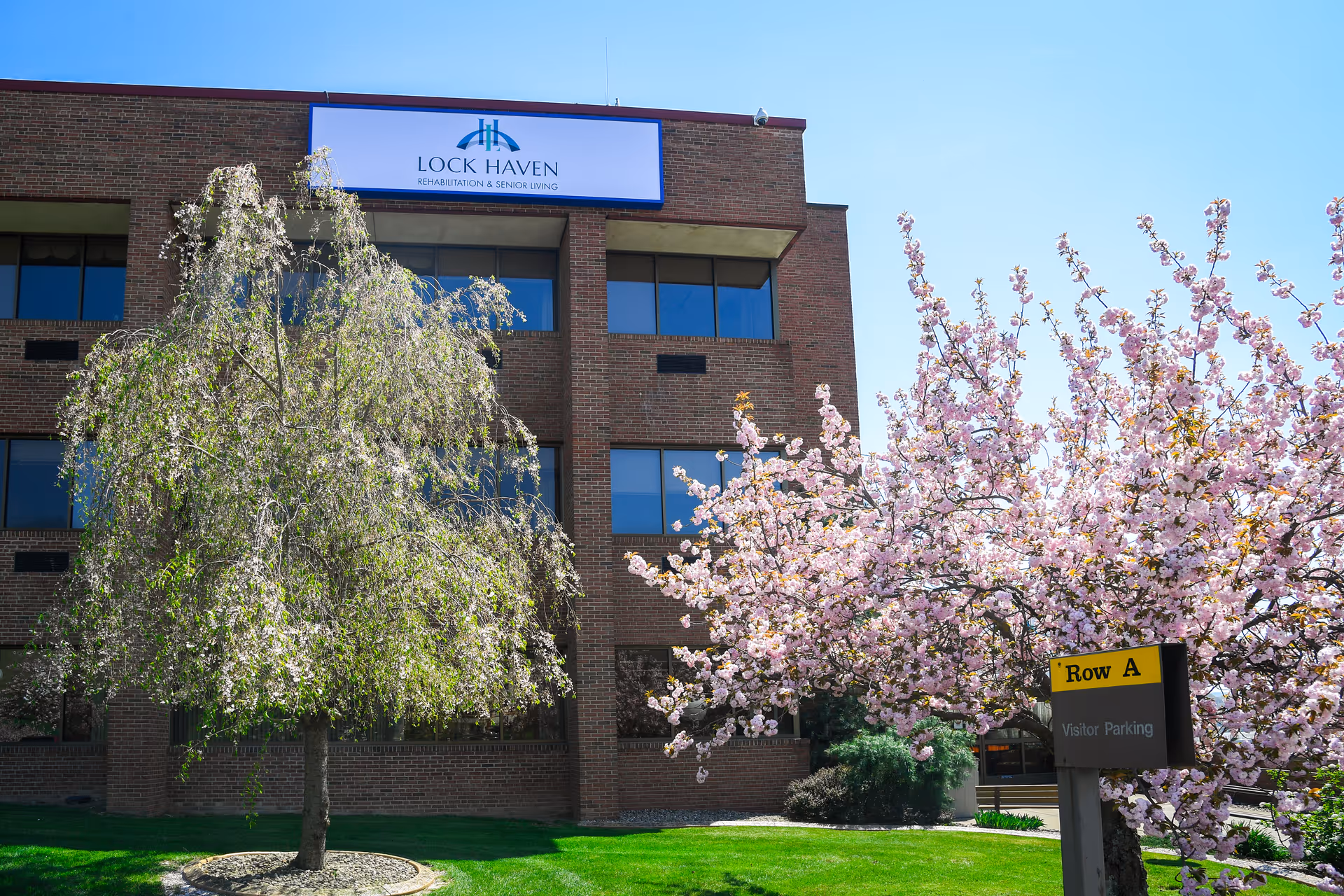 Brick facade of Lock Haven Rehabilitation & Senior Living with flowering trees and a 'Row A Visitor Parking' sign in front.