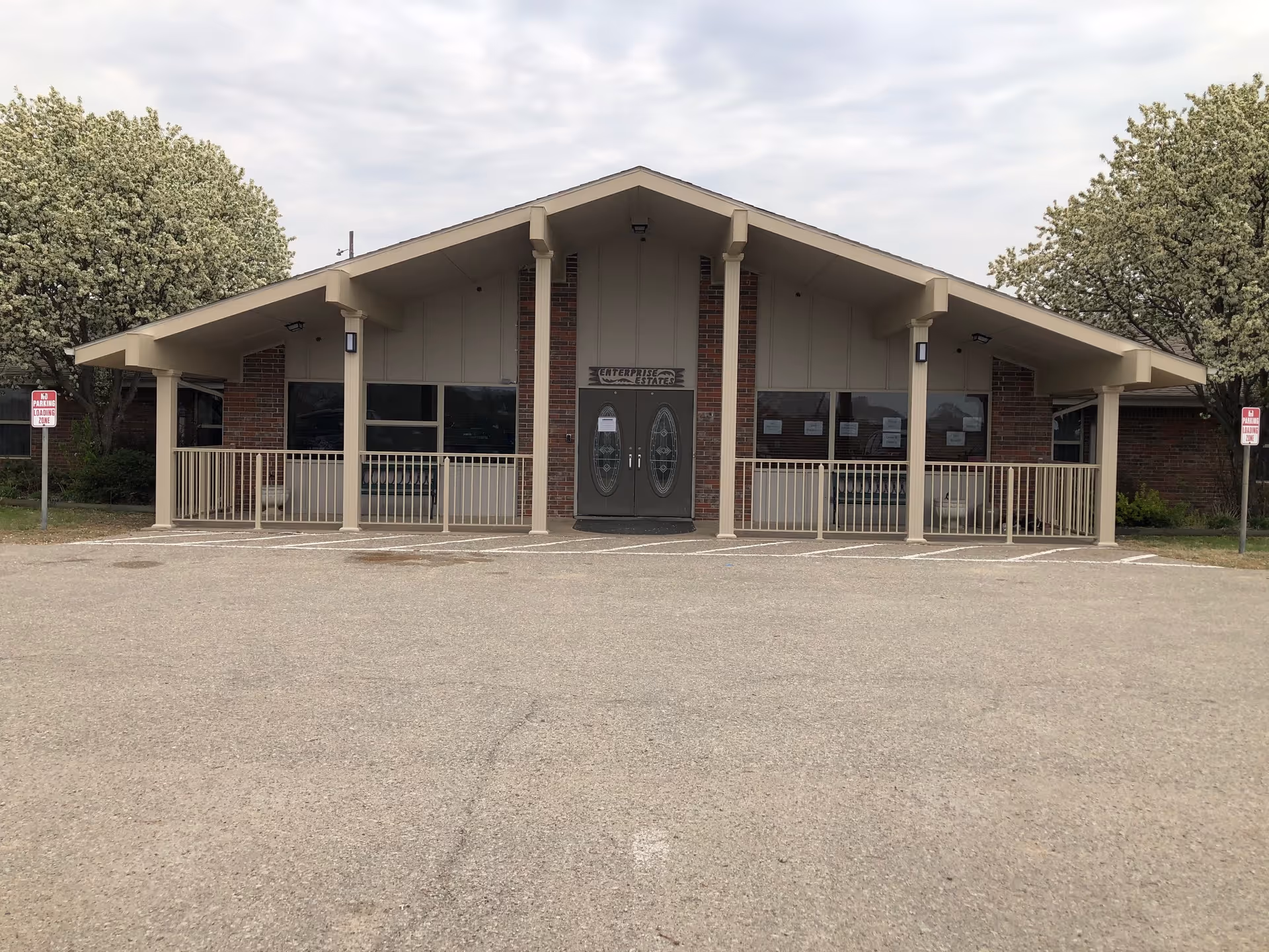 Front exterior view of Enterprise Estates Nursing Center building with a peaked roof, double glass doors, brick and beige siding, two flowering trees on either side, and a parking area in front.