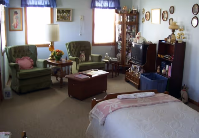 A cozy senior living bedroom with a bed covered in a white quilt and a pink blanket. Two green armchairs with pillows are placed near two windows with blue valances. A wooden coffee table is in front of the chairs, and a wooden cabinet with a TV and various decorative items is against the wall. A small bookshelf and a glass display cabinet with collectibles are also visible.