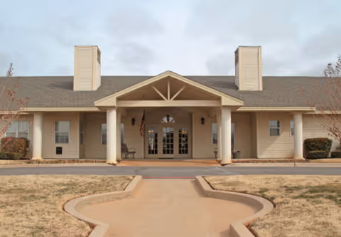 Front entrance of a beige single-story building with a covered portico, double glass doors, and a paved walkway.