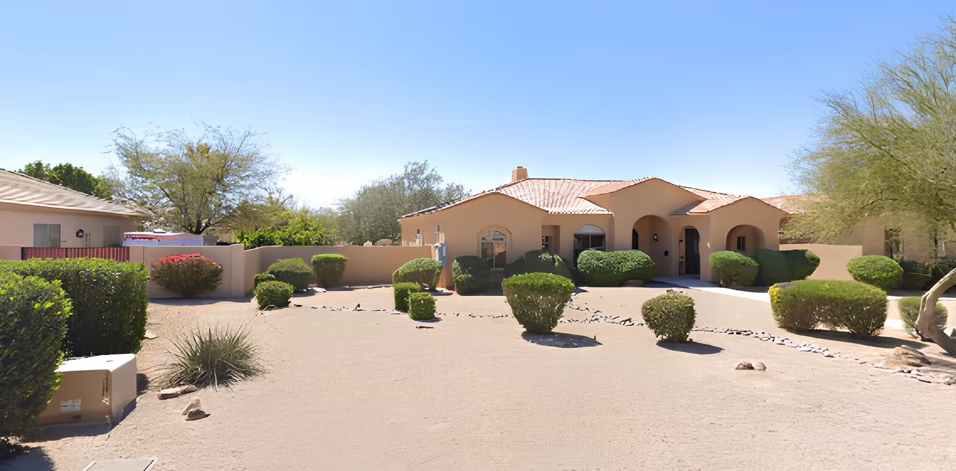 Exterior view of a single-story building with a beige stucco finish and a red-tiled roof, surrounded by desert landscaping including trimmed bushes, small trees, and sandy ground under a clear blue sky.