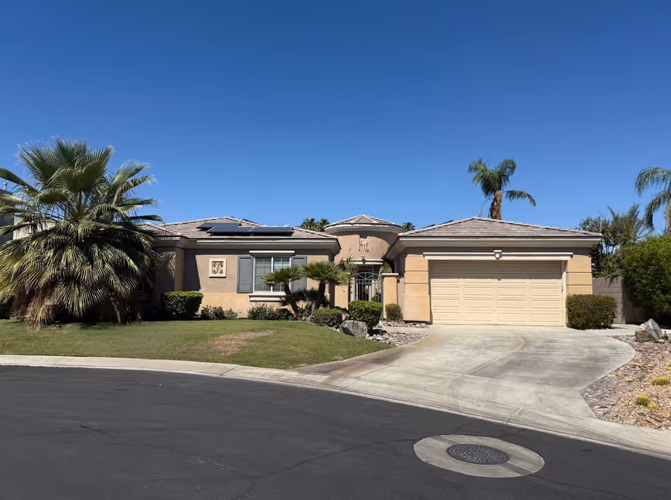 Single-story beige house with a two-car garage, palm trees, and desert landscaping under a clear sky.