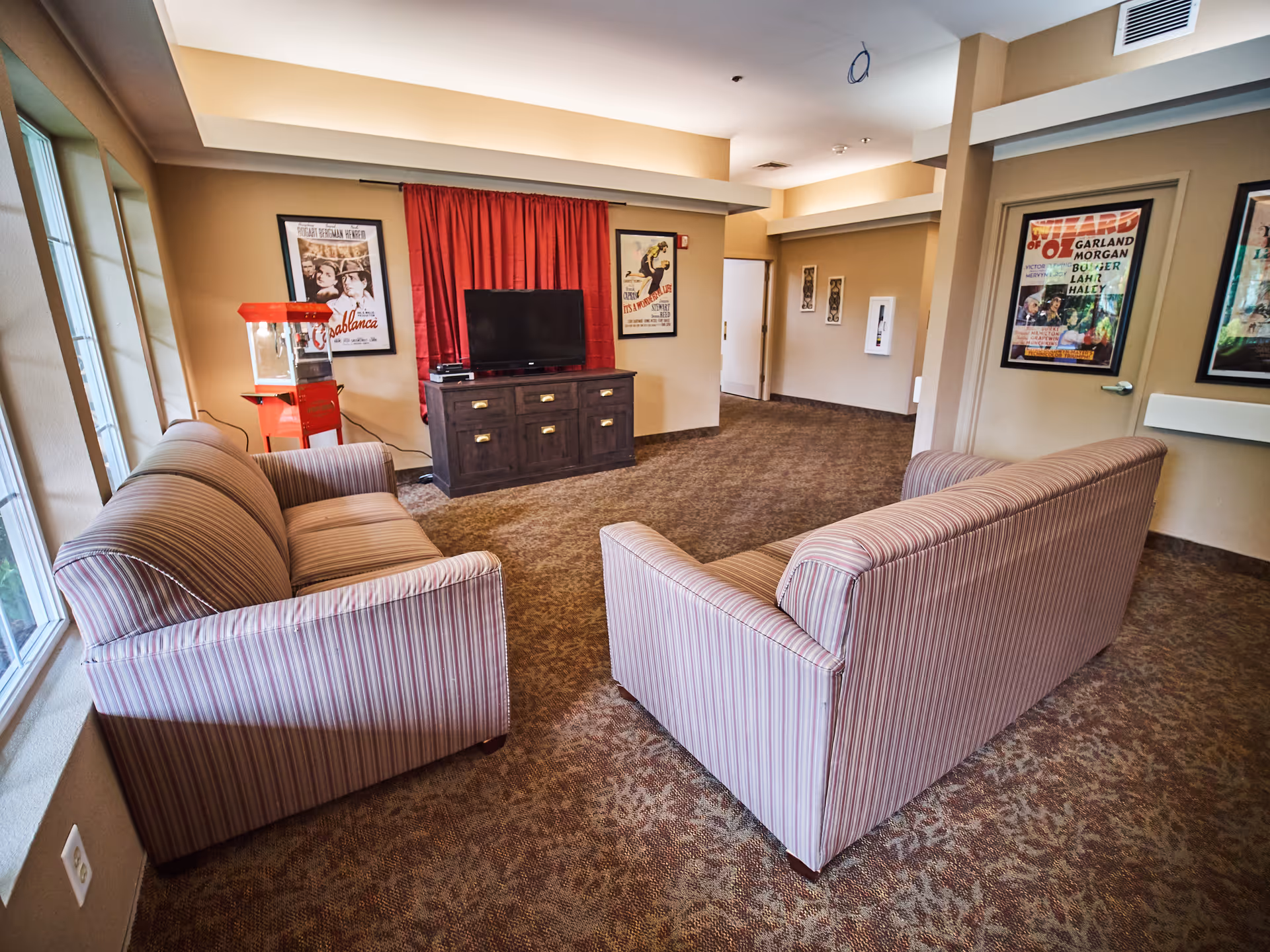 A cozy common area with two striped sofas facing a TV on a wooden cabinet. There is a red popcorn machine next to the TV. The walls are decorated with framed vintage movie posters, and large windows let in natural light from the left side.