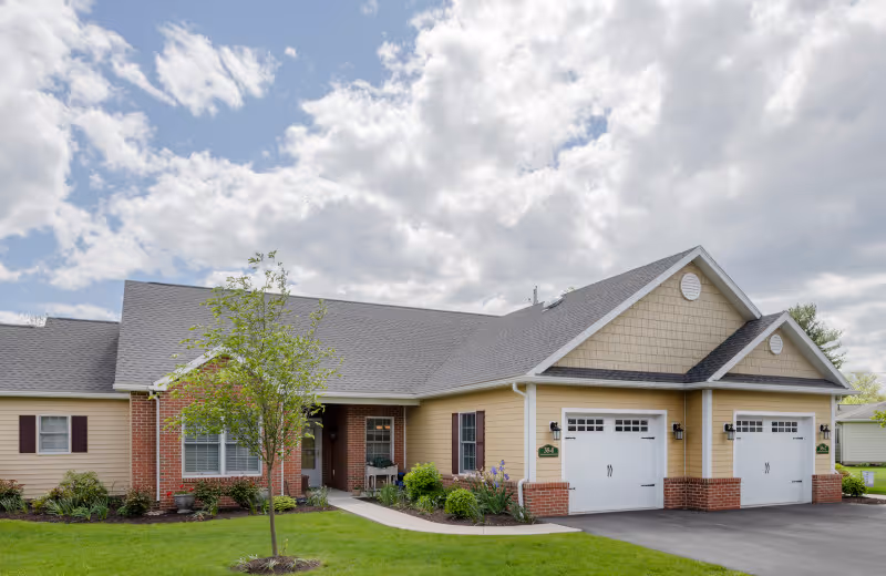Exterior view of a single-story residential building with beige siding and brick accents, featuring two white garage doors, a small tree and landscaped garden in front, under a partly cloudy sky.