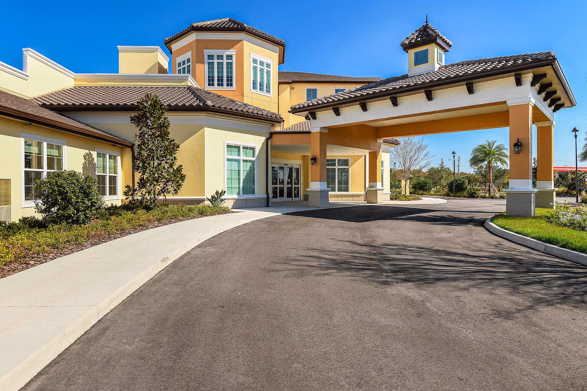 Exterior view of a senior living facility building with a covered entrance driveway, yellow and beige walls, tiled roof, and landscaped greenery under a clear blue sky.