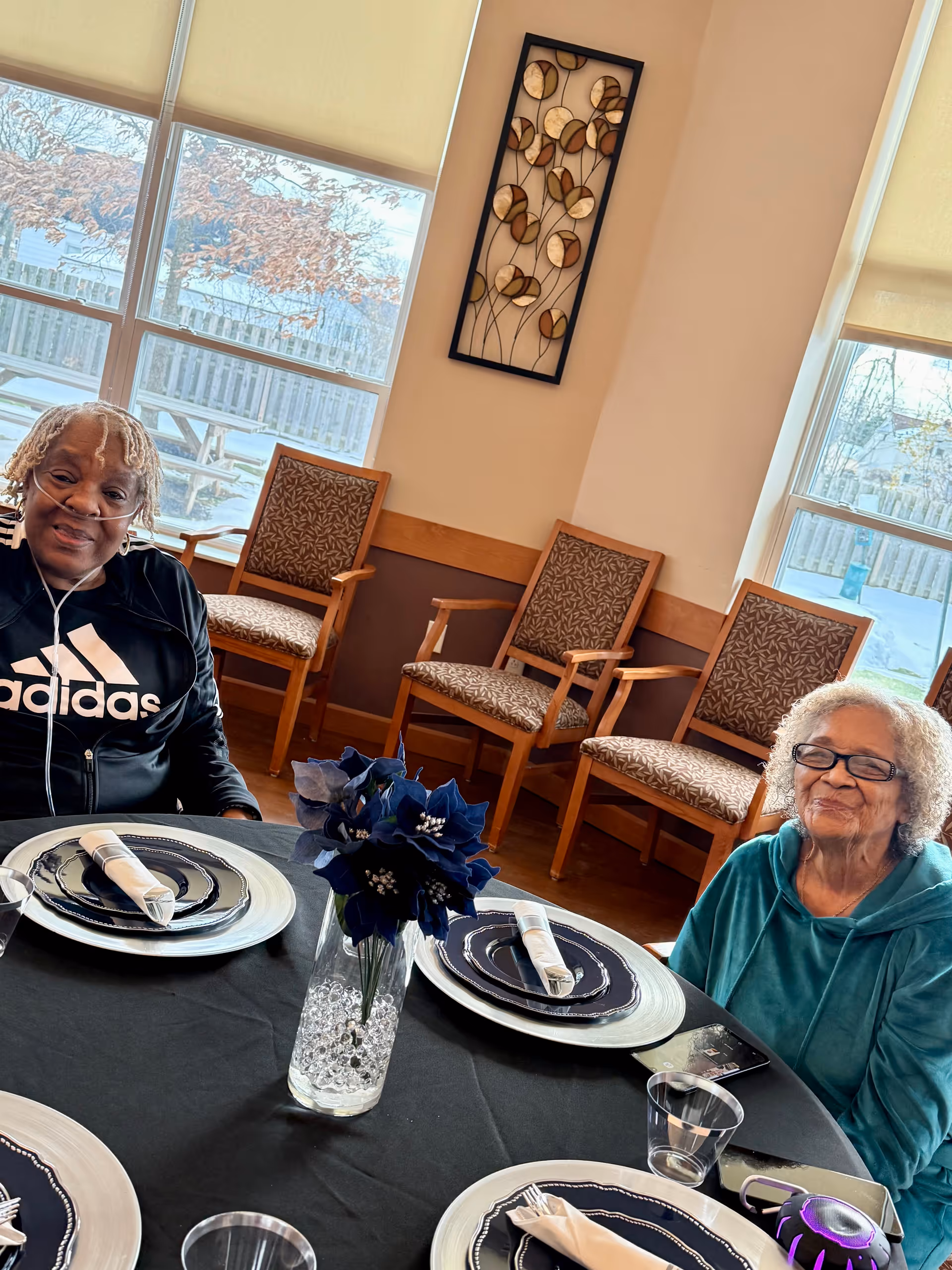 Two seniors sit at a round dining table with place settings and a blue vase centerpiece.