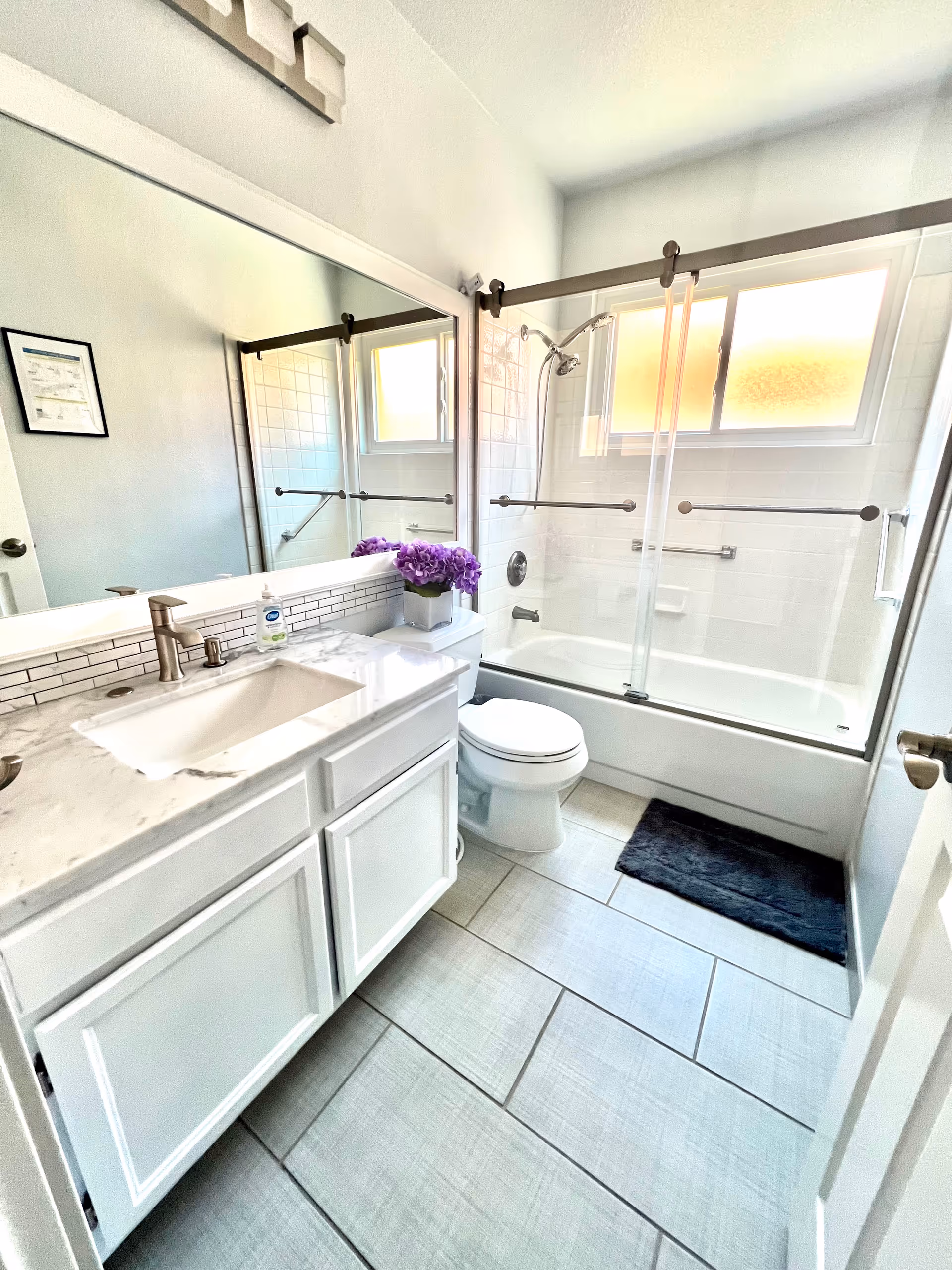 Bright bathroom in a senior living facility with a white vanity, tub/shower, and purple flowers.