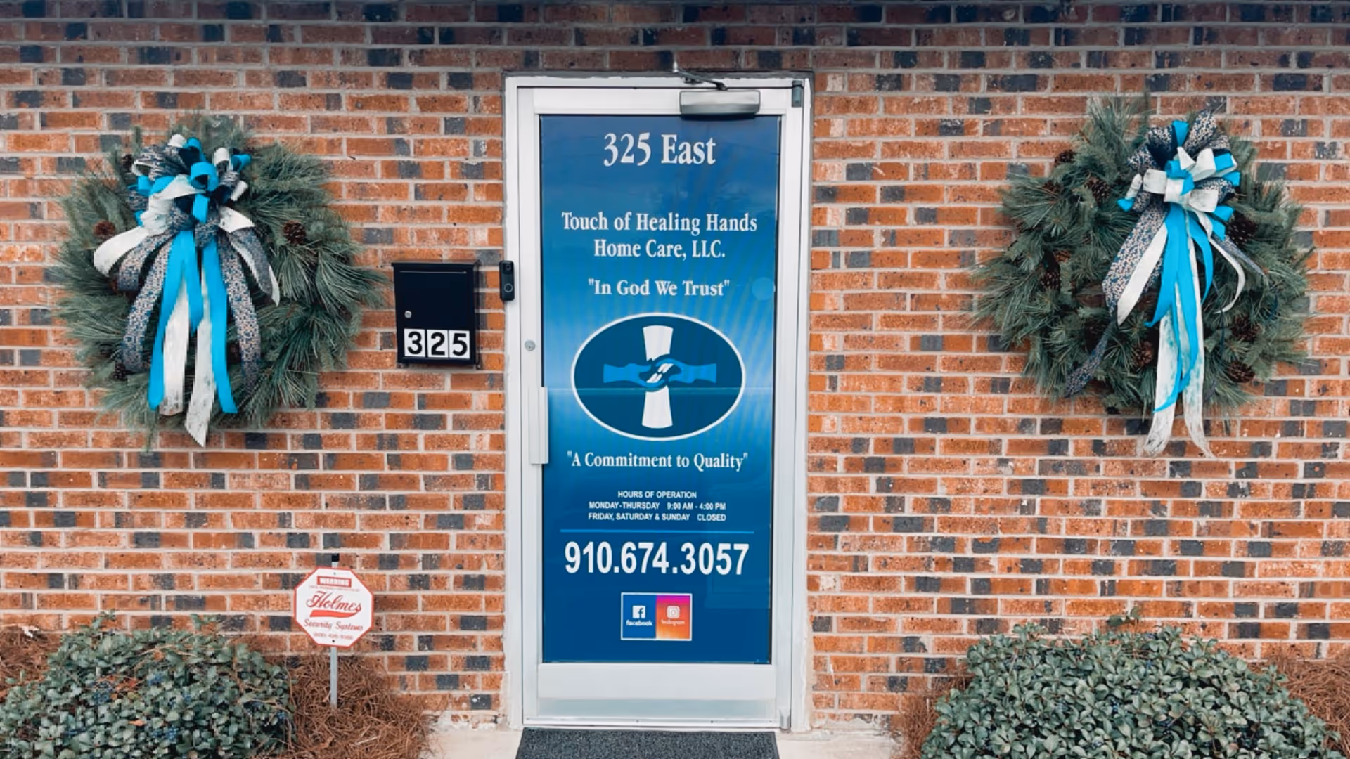 Brick building entrance with blue sign and decorative wreaths beside the door.
