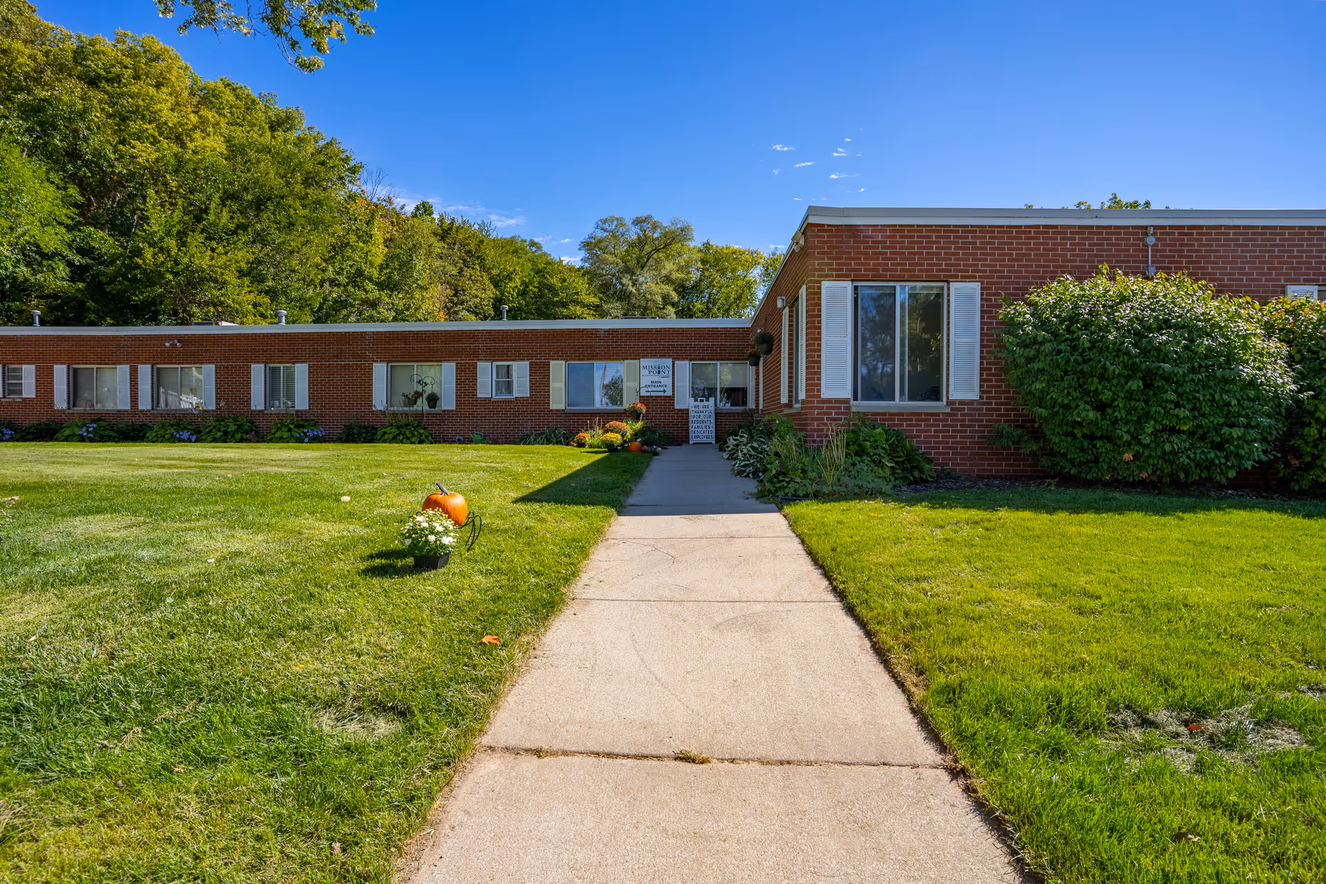 Exterior view of a single-story brick building with white window shutters, surrounded by green grass and bushes under a clear blue sky. A concrete pathway leads to the entrance of the building, which has a sign near the door. Trees are visible in the background.