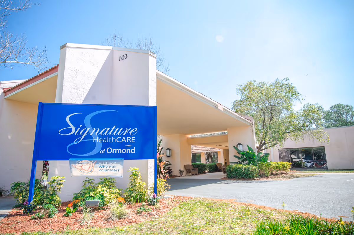 Exterior view of the Signature HealthCARE of Ormond building with a large blue sign in front displaying the facility name. The building has a covered entrance with seating and is surrounded by greenery and trees under a clear blue sky.