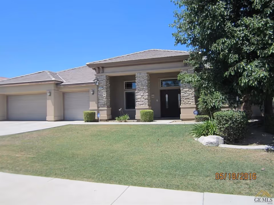 Front exterior view of a single-story house with a three-car garage, stone pillars at the entrance, a well-maintained lawn, and trees on the right side under a clear blue sky.