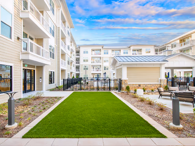 Outdoor courtyard area of a senior living facility with a green lawn, paved walkways, seating areas with chairs and tables, and multi-story residential buildings in the background under a partly cloudy sky.