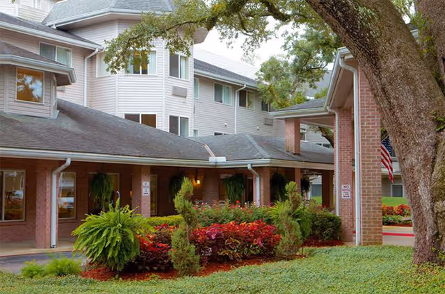 Exterior view of Kemp Meadows Assisted Living facility showing a brick building with multiple windows, a covered entrance, well-maintained landscaping with green shrubs, red flowers, and a large tree in the foreground.