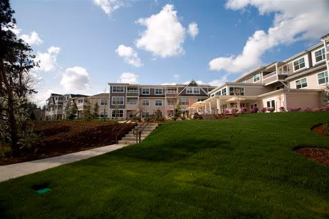 Exterior view of The Stafford Retirement Community building under a partly cloudy sky, with a well-maintained green lawn and a concrete walkway with stairs leading up to the entrance.