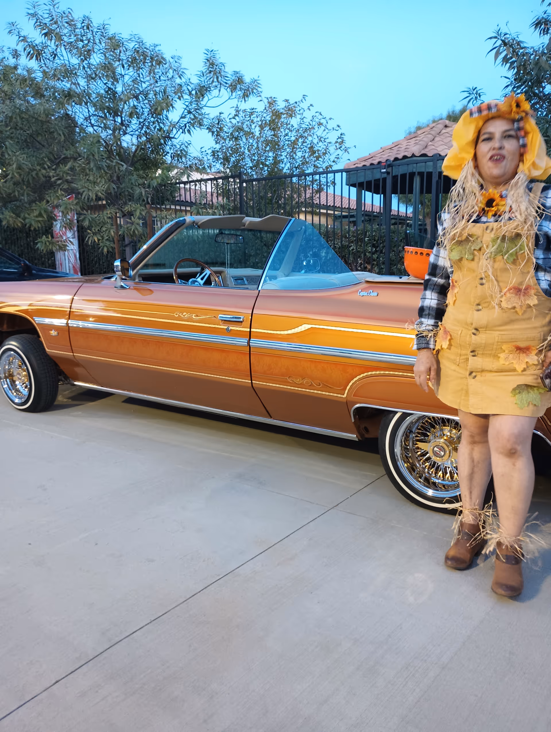 Person in harvest-themed costume stands beside a vintage orange convertible on a driveway.