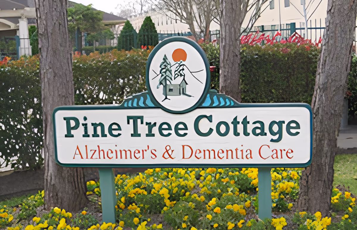 Outdoor sign for Pine Tree Cottage Alzheimer's & Dementia Care surrounded by yellow flowers, trees, and bushes in a garden setting.