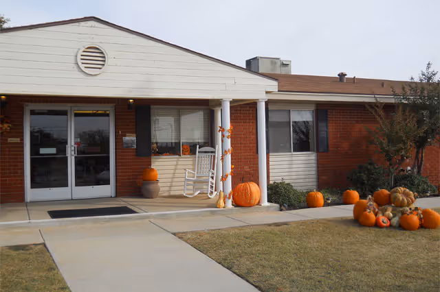 Exterior view of a single-story brick and siding building with a covered entrance. The entrance has double glass doors, a white rocking chair on the porch, and several pumpkins arranged around the porch and lawn, suggesting a fall or Halloween decoration theme.