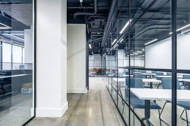 Modern glass-walled corridor leading to a bright dining area with tables and blue chairs.