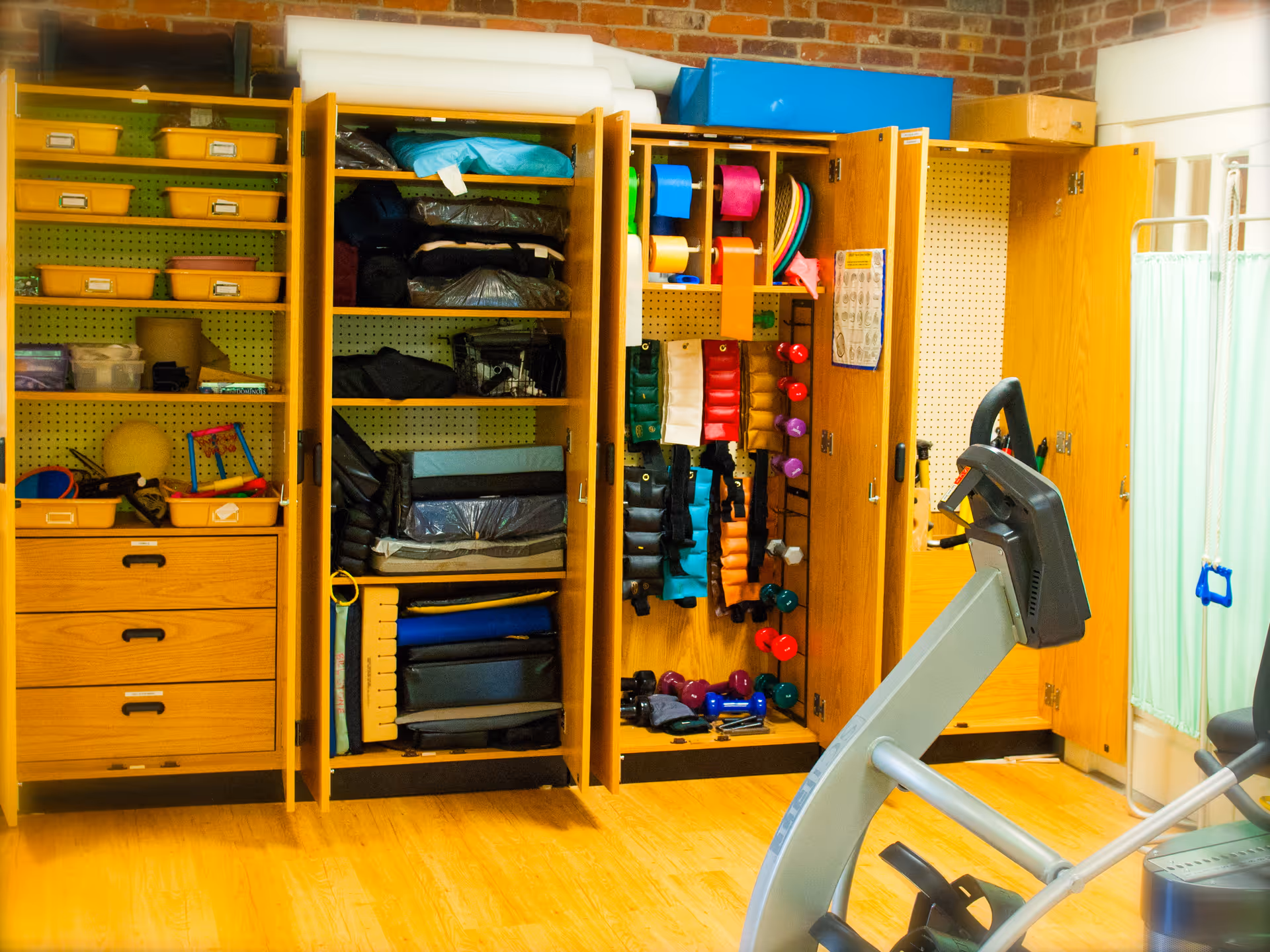 A room with wooden cabinets filled with various exercise equipment including dumbbells, resistance bands, mats, and other fitness accessories. In the foreground, there is a piece of exercise equipment, possibly a stationary bike or elliptical machine. The room has a wooden floor and a brick wall in the background.