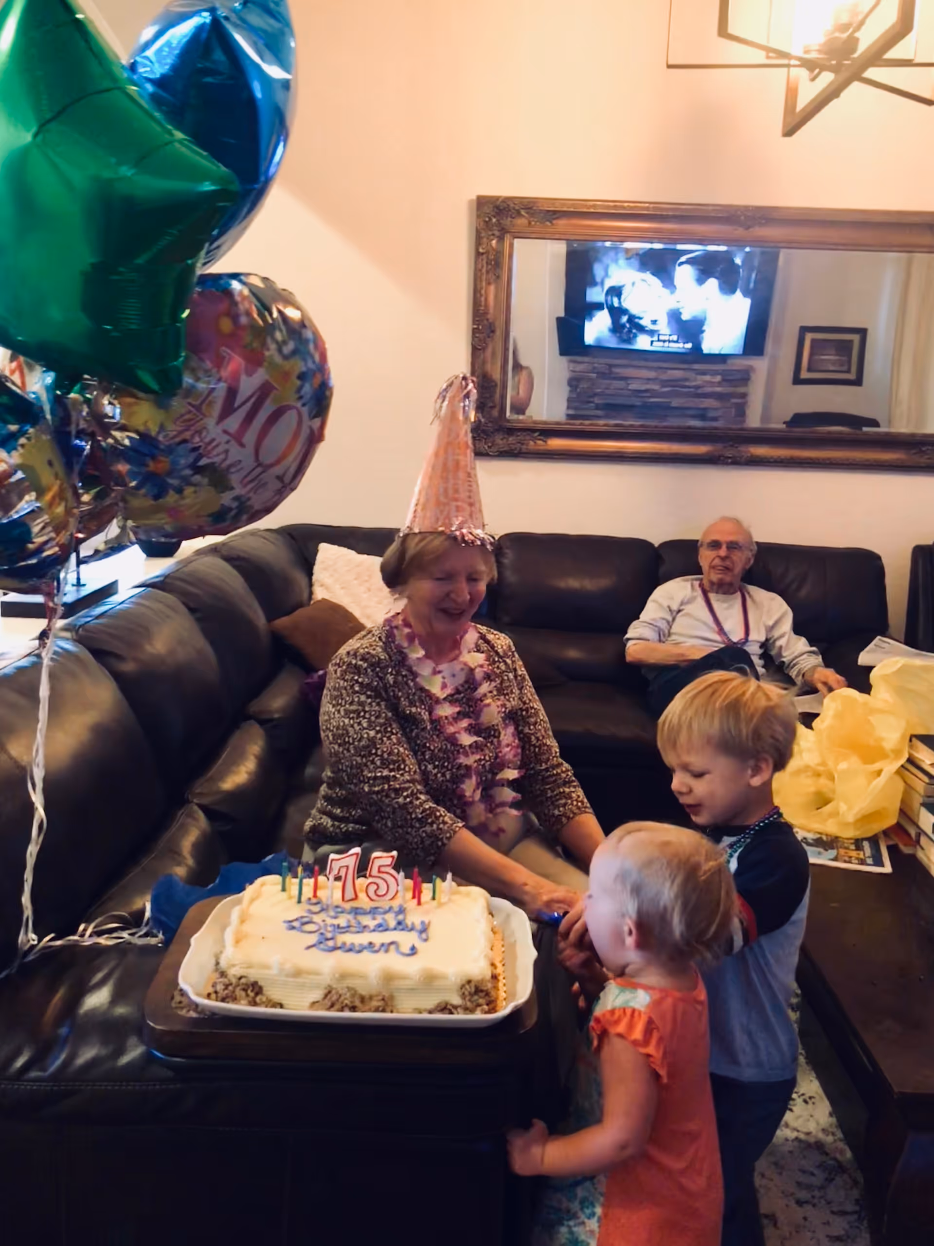 An elderly woman wearing a party hat and a lei is sitting on a dark leather couch, smiling at two young children standing in front of her. A birthday cake with lit candles showing the number 75 is on a table in front of them. Balloons are visible to the left side of the image. An elderly man is sitting on the couch in the background. A large mirror and a television are on the wall behind them.