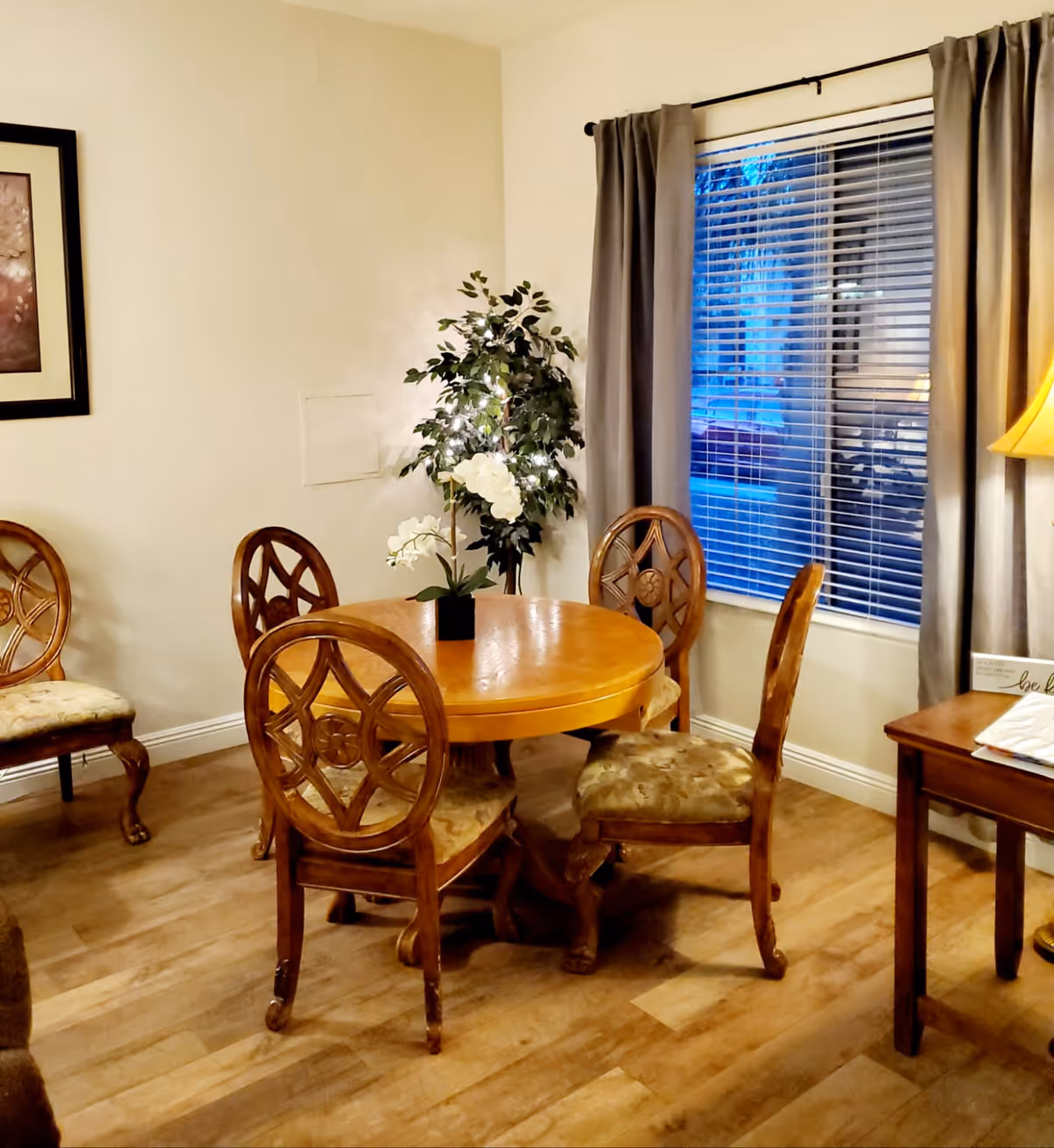 Round wooden dining table with chairs in a cozy room, plant, and window blinds.