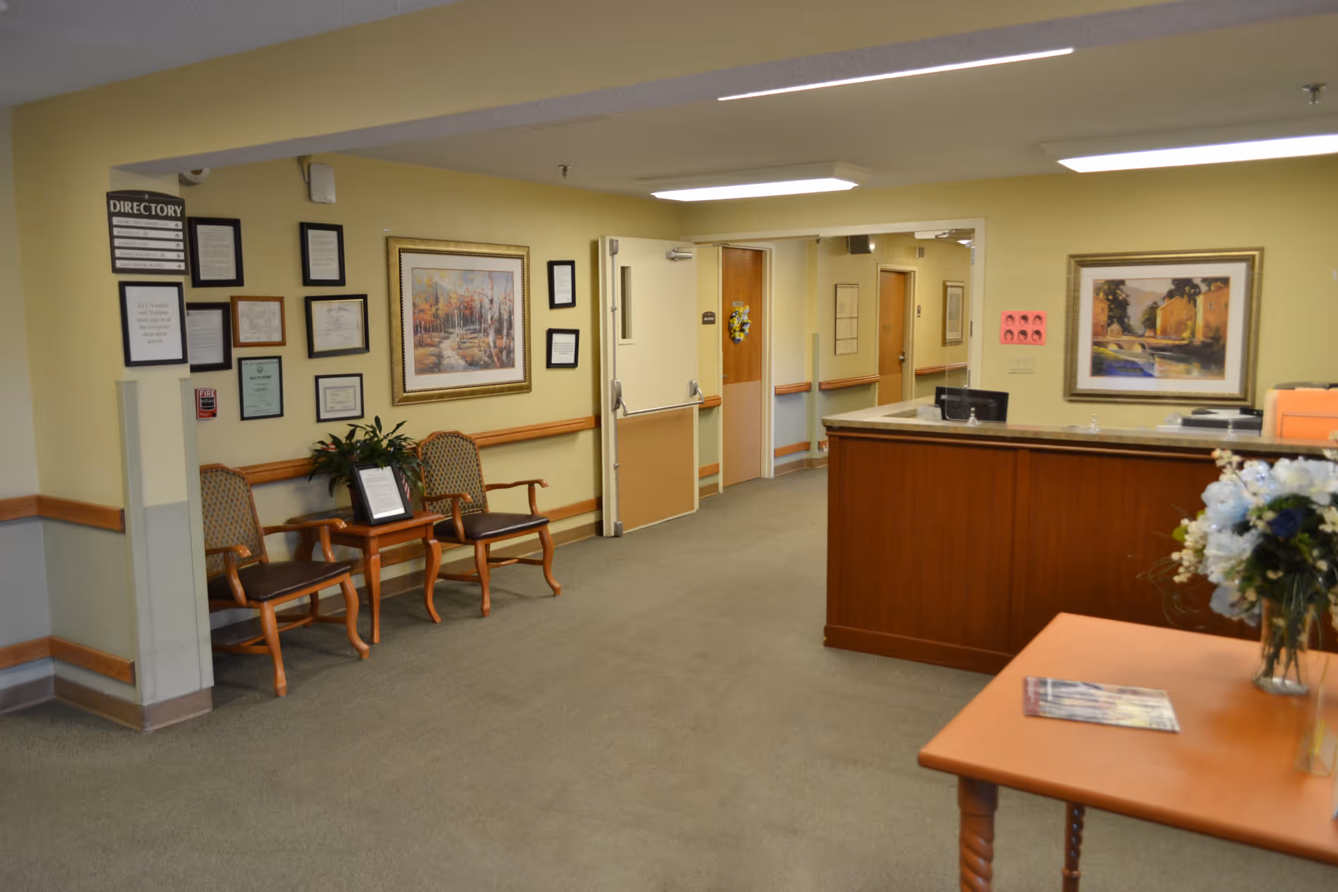 Reception area of a senior living facility with a front desk, seating, framed artwork, and hallways leading deeper into the building.