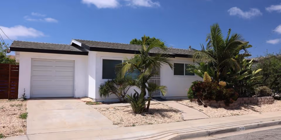 Single-story white house with an attached garage, driveway, and palm trees in front under a blue sky.